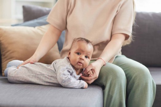 Portrait of cute mixed-race baby looking at camera while crawling on couch with unrecognizable Caucasian mother in background, copy space